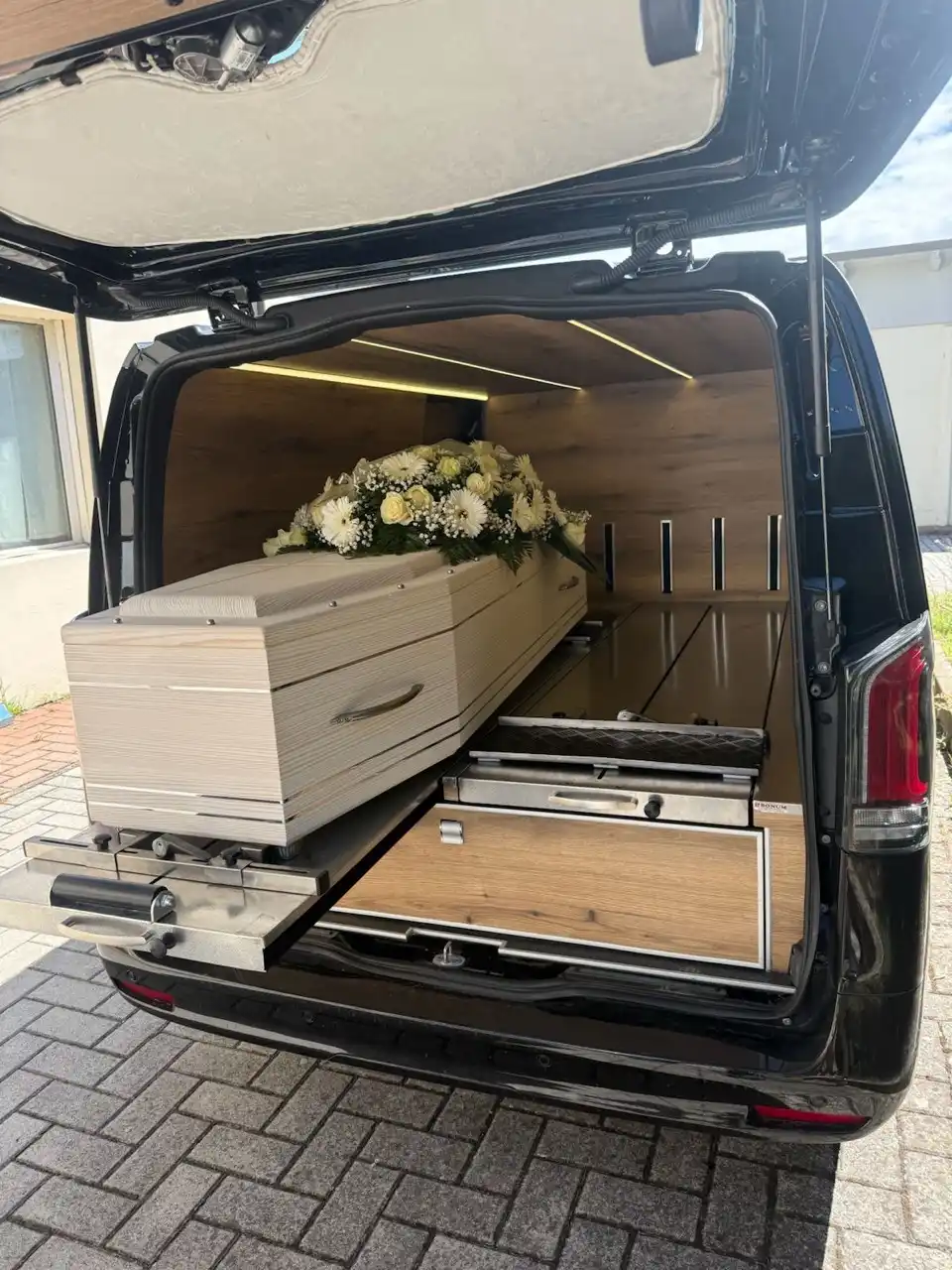 Svetli sanduk sa cvetnim aranžmanom na platformi u otvorenom pogrebnom vozilu.